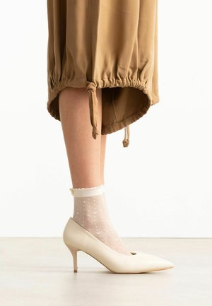 Bas des jambes portant des chaussettes blanches transparentes à pois et des escarpins beige à bout pointu sous une jupe marron à ourlet froncé.