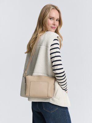 Beige Leder Umhängetasche mit einer Fronttasche, glatter Textur und Reißverschluss, getragen über einer weißen Fleeceweste und gestreiftem Oberteil.