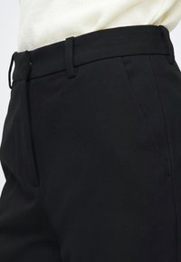 Κοντινή όψη μαύρου tailored παντελονιού με θηλιές για ζώνη και πλαϊνή τσέπη, φορεμένο με λευκό τοπ.