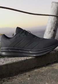 Baskets de sport noires avec côtés texturés et trois bandes, posées sur un rebord en béton devant un arrière-plan extérieur flou.