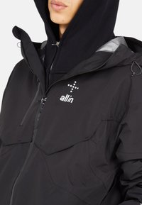 Veste noire imperméable avec capuche, dotée d'une fermeture éclair à l'avant, de deux poches poitrine et d'un logo blanc sur le côté gauche. Texture lisse, design minimaliste.
