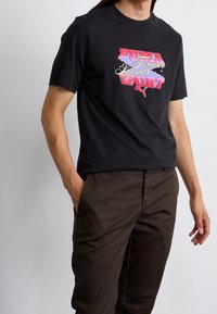 T-shirt in cotone nero con una stampa grafica colorata e il testo "PUMA Forever Legends Sport" insieme a un logo, abbinato a pantaloni marroni.