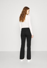 Calças flared pretas com um acabamento texturizado, apresentando uma cintura média e detalhes nos bolsos traseiros, combinadas com uma blusa de manga longa branca.