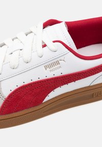 Baltas ādas kedas ar sarkaniem zamšiem akcentiem, baltām auklām, gumijas zoli un perforētiem detaļām. Iespējo "PUMA INDOOR" zīmolu.