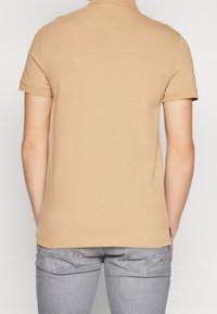 Beżowa koszulka polo z krótkim rękawem o miękkiej fakturze, z płaskim kołnierzykiem i bocznymi rozporkami, zestawiona z jasnoszarymi dżinsami. Widok z tyłu ukazuje estetyczne linie.