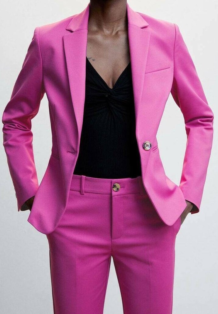 Femme en tailleur rose vif sur mesure avec veste et pantalon, portant un top noir torsadé sur le devant en dessous, mains dans les poches.