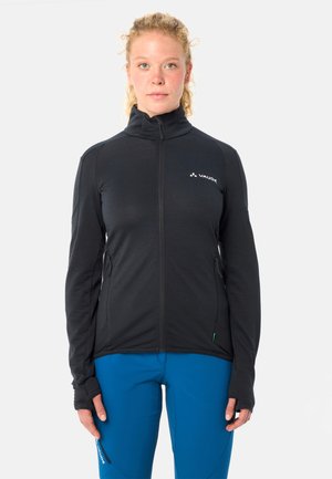 Vrouw met krullend blond haar, gekleed in een zwart Vaude-jas met ritssluiting en blauwe broek, staand tegen een effen lichte achtergrond.