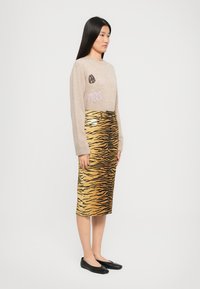 Femme aux longs cheveux noirs portant un pull beige avec des patchs et une jupe mi-longue à rayures tigre jaunes, accompagnée de ballerines noires.