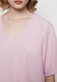 Top rosa chiaro a V con maniche corte e vestibilità rilassata, caratterizzato da una texture liscia e un sottile dettaglio di cucitura al centro.