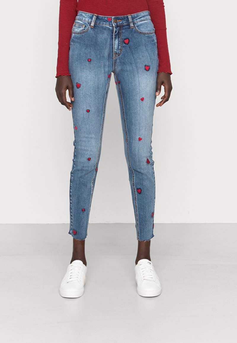 Femme portant un jean skinny bleu avec des patchs en forme de cœur rouges, des baskets blanches et un haut rouge à manches longues, debout sur un sol gris clair.
