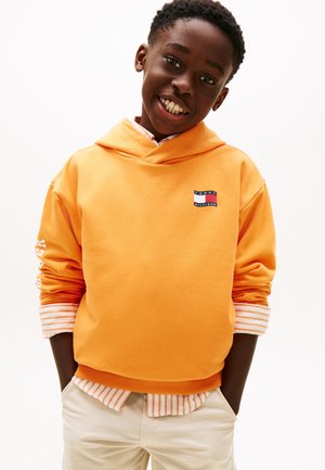 Băiat zâmbitor purtând o hanorac portocaliu Tommy Hilfiger peste un tricou în dungi, cu mâinile în buzunarele pantalonilor bej, pe un fundal alb.