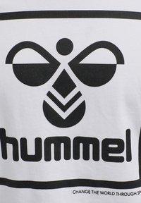 Camiseta blanca con un destacado diseño gráfico negro que presenta formas abstractas y el nombre de la marca "hummel" en letras negritas en la parte inferior.