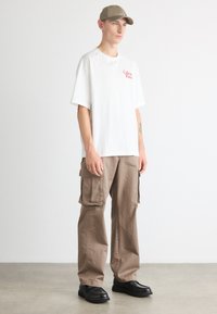 Hvid t-shirt med rødt logo, parret med tan cargo-bukser med store lommer og sorte snørebåndssko. Modellen bærer en lys cap.