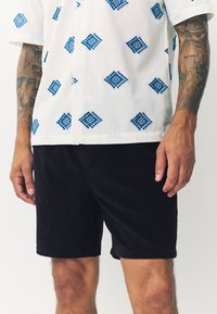 Witte shirt met korte mouwen en blauwe ruitpatronen, gecombineerd met zwarte corduroy shorts. Close-up van handen die op de shorts rusten.