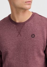 Bordeaux sweatshirt lavet af blødt stof, med et ribbet rundhalsudskæring, skulderdetaljer og et lille sort logo på brystet.