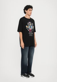 Black oversized t-shirt with colorful graphic print, featuring butterflies and text. Paired with dark blue jeans and black sneakers.