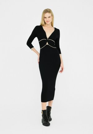 Une modèle pose dans une robe noire ajustée avec un décolleté en V profond et des accents dorés, associée à des bottines noires, sur un fond blanc uni.