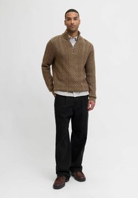 Brauner Kabelstrickpullover mit hohem Kragen und Reißverschlussdetails, getragen über einem weißen Hemd. Kombiniert mit schwarzen Hosen und braunen Schuhen.