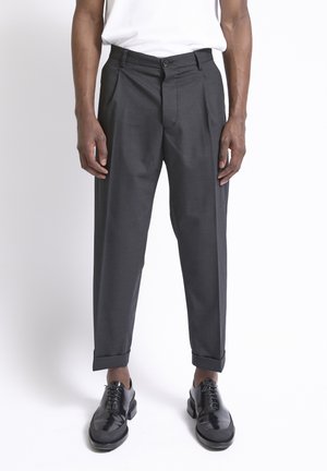 Uomo con pantaloni grigi scuri risvoltati, scarpe eleganti nere in pelle e camicia bianca infilata, in piedi davanti a uno sfondo neutro.