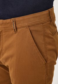 Braune Baumwollhosen mit glatter Textur, ausgestattet mit Taschen und einem Reißverschluss, teilweise sichtbar gegen einen dunklen Strickstoff.