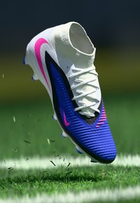 Ποδοσφαιρικό παπούτσι Nike με λευκό γιακά, μπλε και μαύρο σώμα, ροζ λεπτομέρειες, ραβδωτή υφή και πολύχρωμες τρύπες, να αιωρείται πάνω από το γρασίδι.