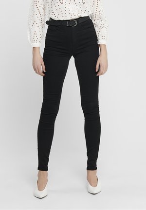 Femme portant un pantalon skinny taille haute noir avec une ceinture noire, un chemisier blanc à œillets et des talons blancs à bout pointu, debout devant un fond uni.