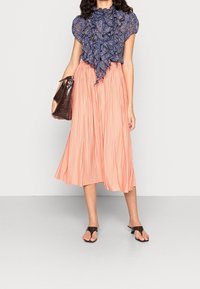 Blusa blu a stampa con frontale arricciato, abbinata a una gonna midi a pieghe color pesca. Una borsa in finta pelle di coccodrillo marrone e sandali neri completano il look.