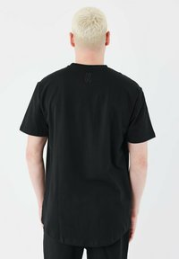 Camiseta de algodón negra con mangas cortas, escote redondo y dobladillo recto. Presenta una "K" bordada cerca del cuello en la parte posterior.