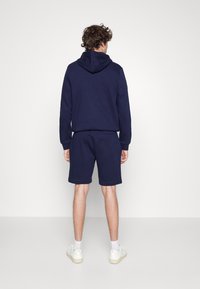 Homme portant un sweat à capuche bleu marine et un short assorti, debout face à un mur blanc uni avec des baskets blanches et des chaussettes.