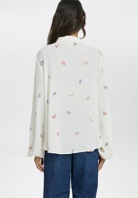 Blusa bianca con maniche lunghe, caratterizzata da ricami floreali colorati. Tessuto leggero, vestibilità comoda, orlo curvo, indossata sopra jeans in denim blu.