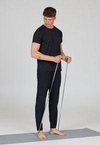Man in zwarte sportkleding staat op een yogamat, gebruikt een weerstandsband voor oefeningen en kijkt naar zijn handen.