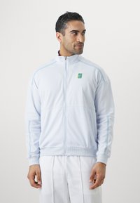Gaiši zila sporta jaka ar rāvējslēdzēja priekšpusi, ribotiem piedurkņu galiem un smalkiem ziliem akcentiem. Uz krūtīm ir zaļš logotips.