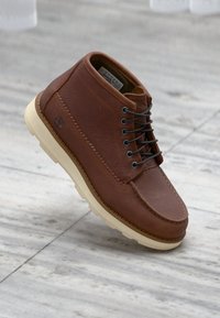 Καφέ δερμάτινο μποτάκι αστραγάλου με ραμμένα διακοσμητικά, στρογγυλή μύτη, μαύρα κορδόνια και λαστιχένια σόλα σε κρεμ απόχρωση. Φέρει λογότυπο στο πλευρό.