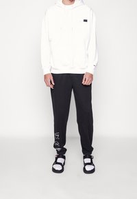 Felpa bianca con cappuccio e tasca frontale, pantaloni sportivi neri con testo grafico bianco e sandali neri con cinturini bianchi e fibbie regolabili.