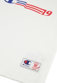 Camiseta blanca de algodón con un gráfico del logo de Champion en azul y rojo, y una etiqueta con texto rojo, situada en la esquina inferior derecha.