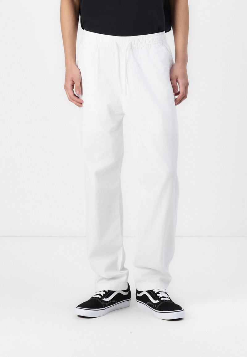 Pantaloni bianchi, leggeri, con vita elasticizzata, dal fit rilassato e gambe dritte, abbinati a sneakers nere Vans con accenti bianchi.