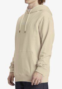 Felpa beige con cappuccio regolabile, tasca frontale, polsini e orlo a coste, texture liscia, con dettaglio logo discreto sul petto.