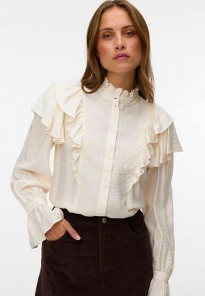 Femme portant un chemisier crème à volants avec boutons sur le devant et col montant, associé à un pantalon en velours côtelé foncé taille haute.