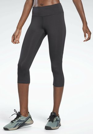 Czarne legginsy o slim-fit, krótkie, noszone przez stojącą osobę, zestawione z szaro-czarnymi butami sportowymi na jednolitym tle.