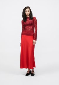 Rdeča dolgoročna majica iz čipke preko gladke, rdeče satenaste maksi krila. Model nosi črne čevlje z ostro konico in stoji pred belim ozadjem.