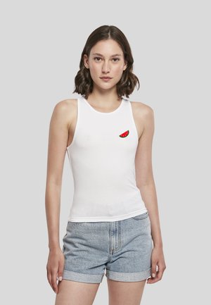 Femme aux cheveux bruns courts portant un haut blanc sans manches avec un petit motif pastèque et un short en jean bleu clair.