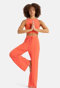 Oranžový rebrovaný crop top a zodpovedajúce široké nohavice s šnúrkou v páse. Modelka v jogovom postoji na jednej nohe, bosými nohami.