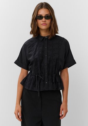 Femme portant un chemisier noir à manches courtes boutonné avec un cordon à la taille et des lunettes de soleil noires sur un fond gris clair.
