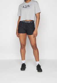 Camiseta gris corta con el logo de "Under Armour"; pantalones cortos deportivos negros con cintura elástica que presentan letras blancas; combinados con zapatillas negras.