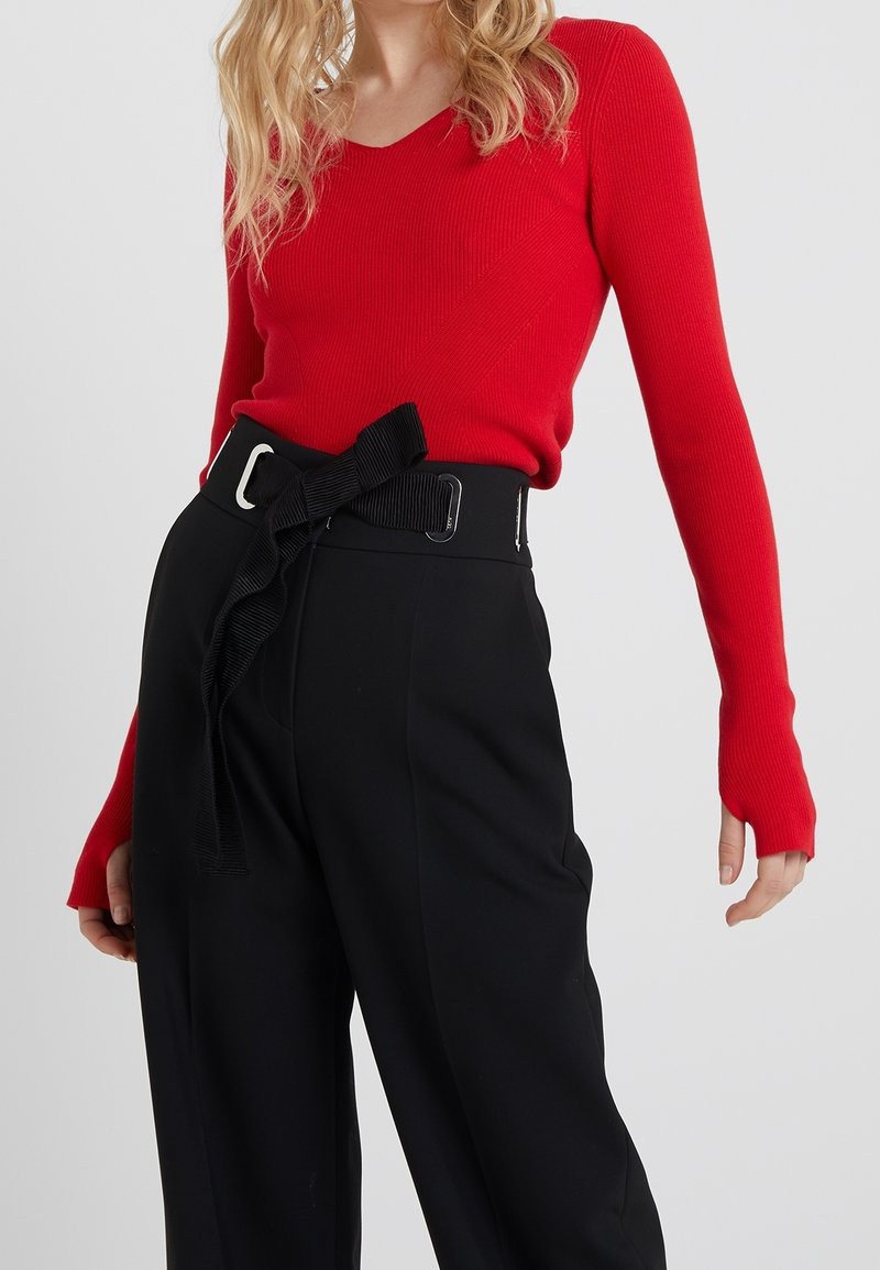 Femme portant un haut rouge ajusté à manches longues, rentré dans un pantalon noir taille haute avec une large ceinture nouée en un nœud.