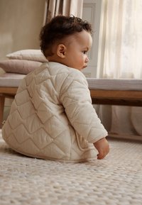 Veste matelassée beige à motif en zigzag, portée par un enfant assis sur un tapis texturé de couleur claire. Tissu doux, col rond et manches longues.