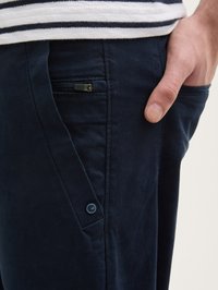 Chino bleu marine avec une finition texturée, dotés d'une poche zippée sur le côté et d'un petit détail de bouton de marque. Coupe et design décontractés.