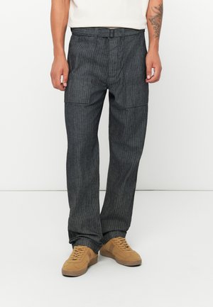 Hombre vestido con pantalones de rayas grises oscuros de corte holgado con grandes bolsillos frontales y zapatillas de ante beige, de pie contra un fondo blanco.