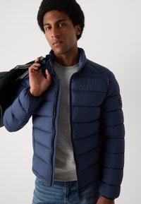 Doudoune marine avec un design matelassé horizontal, fermeture éclair, et col montant, portée sur un t-shirt gris. Sac noir porté en bandoulière.