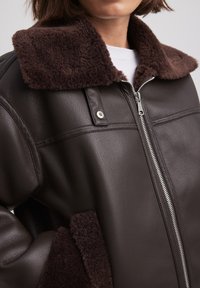 Veste en cuir marron avec un col en shearling souple brun foncé, fermeture éclair argentée et épaules structurées avec des coutures définies.